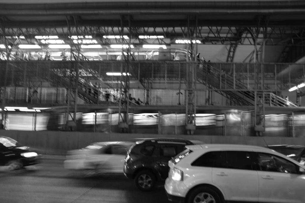 A blurred black and white image showing cars traveling down a highway at night. There is a parking garage in the background with pedestrians descending two stairways.